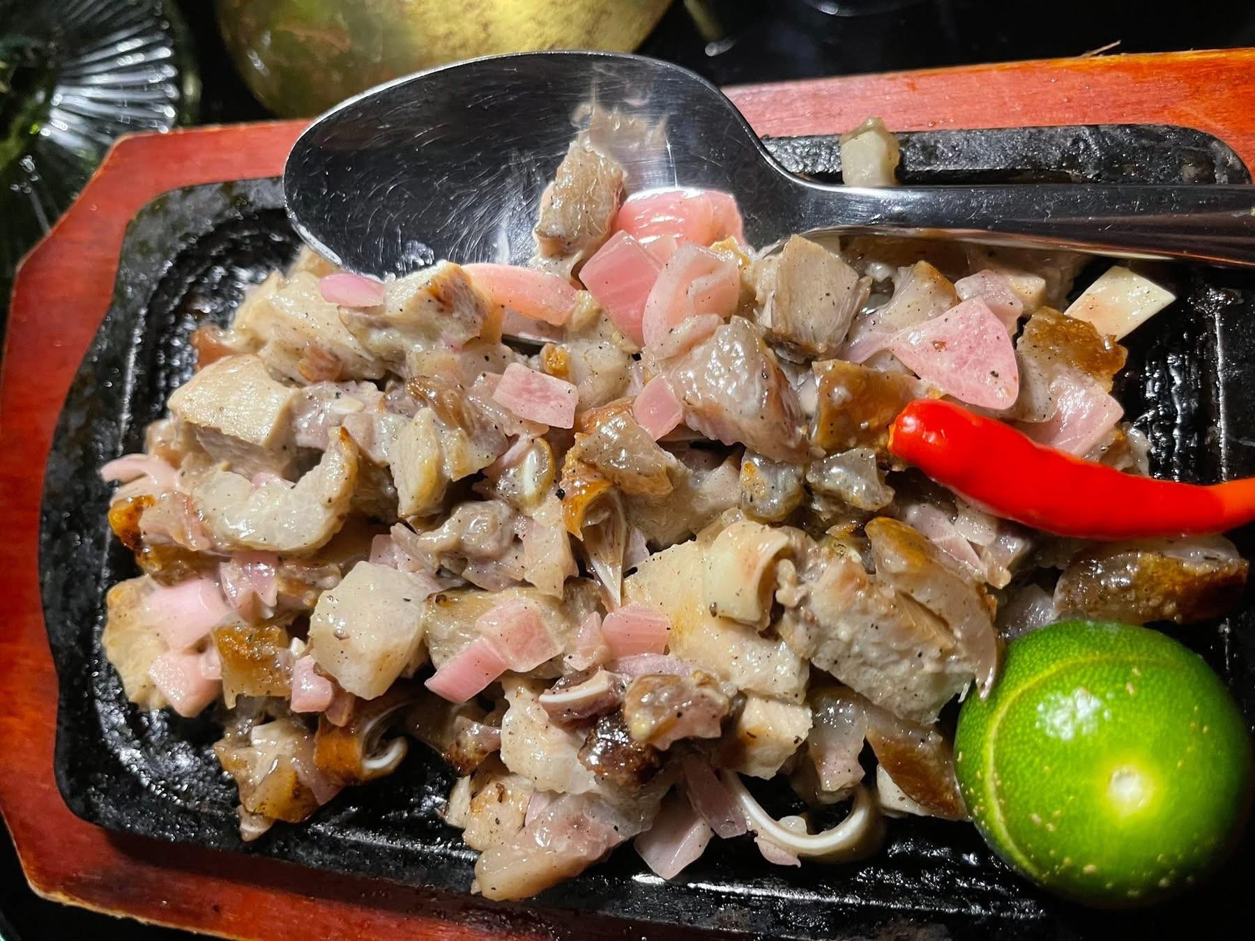 這是一張菲律賓經典料理 Sisig（鐵板碎豬肉）的特寫照片。在黑色的鑄鐵盤上，盛裝著切成小塊的豬頭皮與豬肉，混合著紫洋蔥碎，呈現出豐富的油脂光澤與酥脆質感。盤子右側擺放著一顆鮮綠色的金桔（Calamansi）與一根紅辣椒作為提味佐料，一支金屬湯匙隨意地放置在料理上方，暗示著即將攪拌食用的動態感。