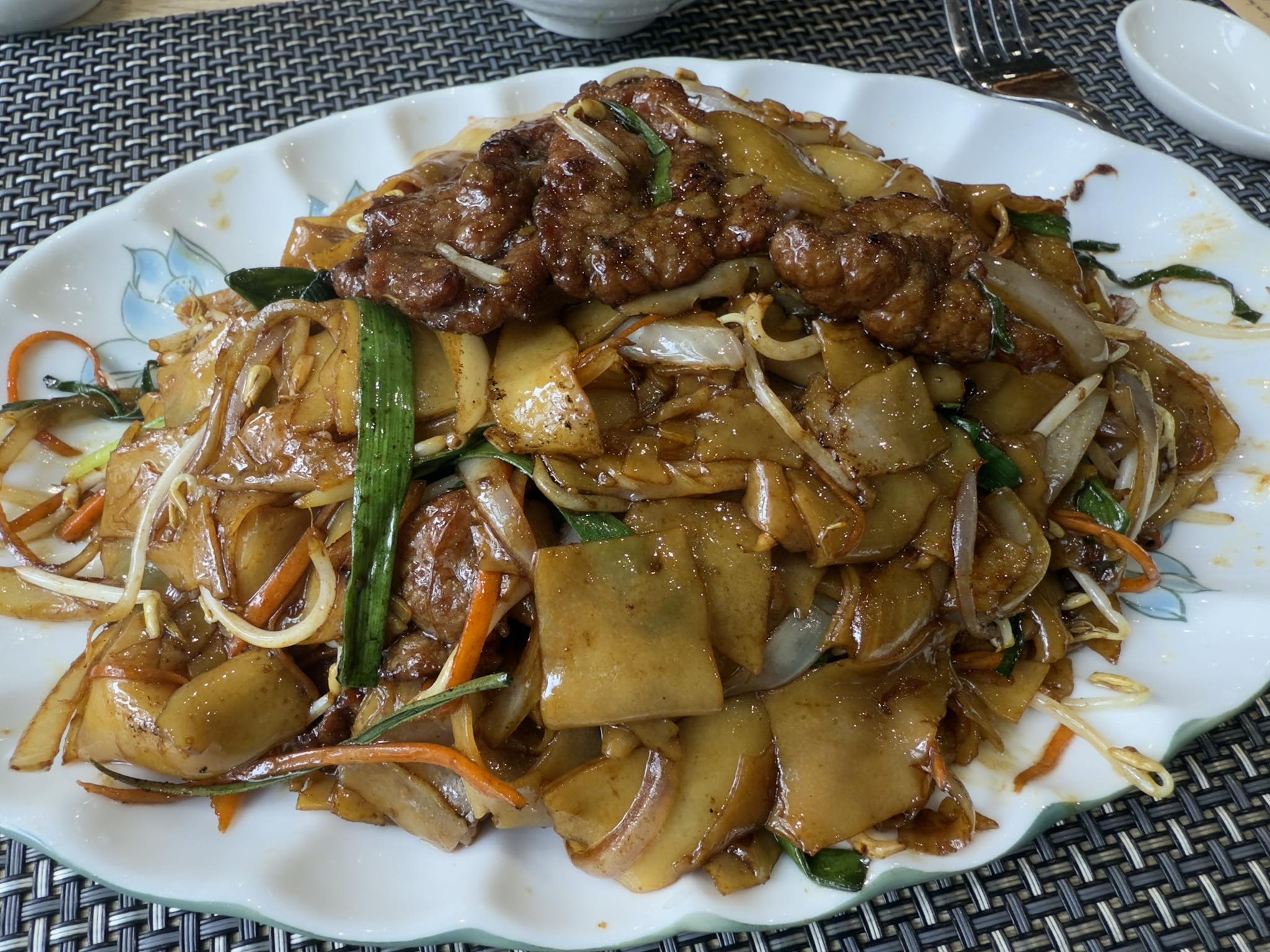 河內街頭餐館拍攝的炒河粉實景：寬扁米麵以大火鑊炒，拌入豆芽、韭菜、洋蔥與紅蘿蔔絲，上覆甜鹹醬燒牛肉與醬油光澤，呈現出廣府沙河粉隨潮汕與廣府移民南漂後，在魚露與在地調味滲入下形成的南洋庶民料理樣貌；我在這盤熱氣蒸騰的河粉裡，看見一條橫跨華南與東南亞的飲食文化遷徙與集體記憶軌跡。