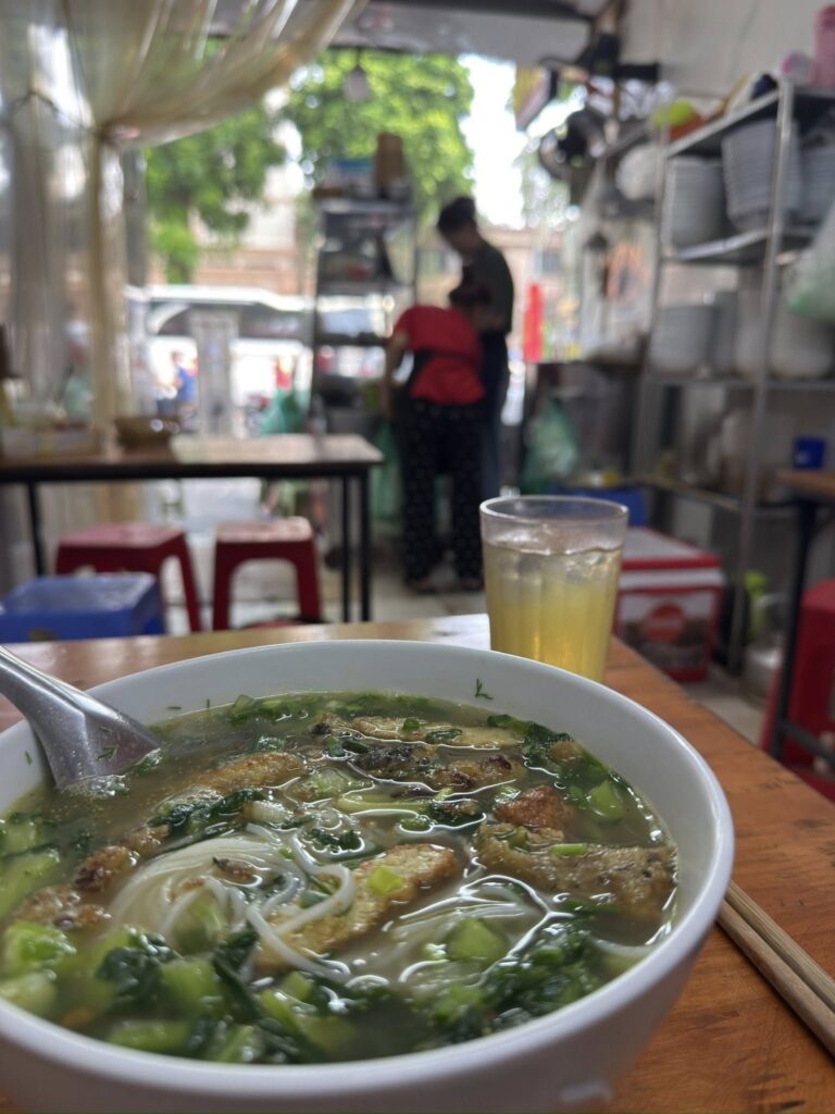 河內街邊小店裡的一碗興安魚肉河粉，前景是熱騰騰的魚粉與冰茶，背景是店家在檯前忙碌的身影。