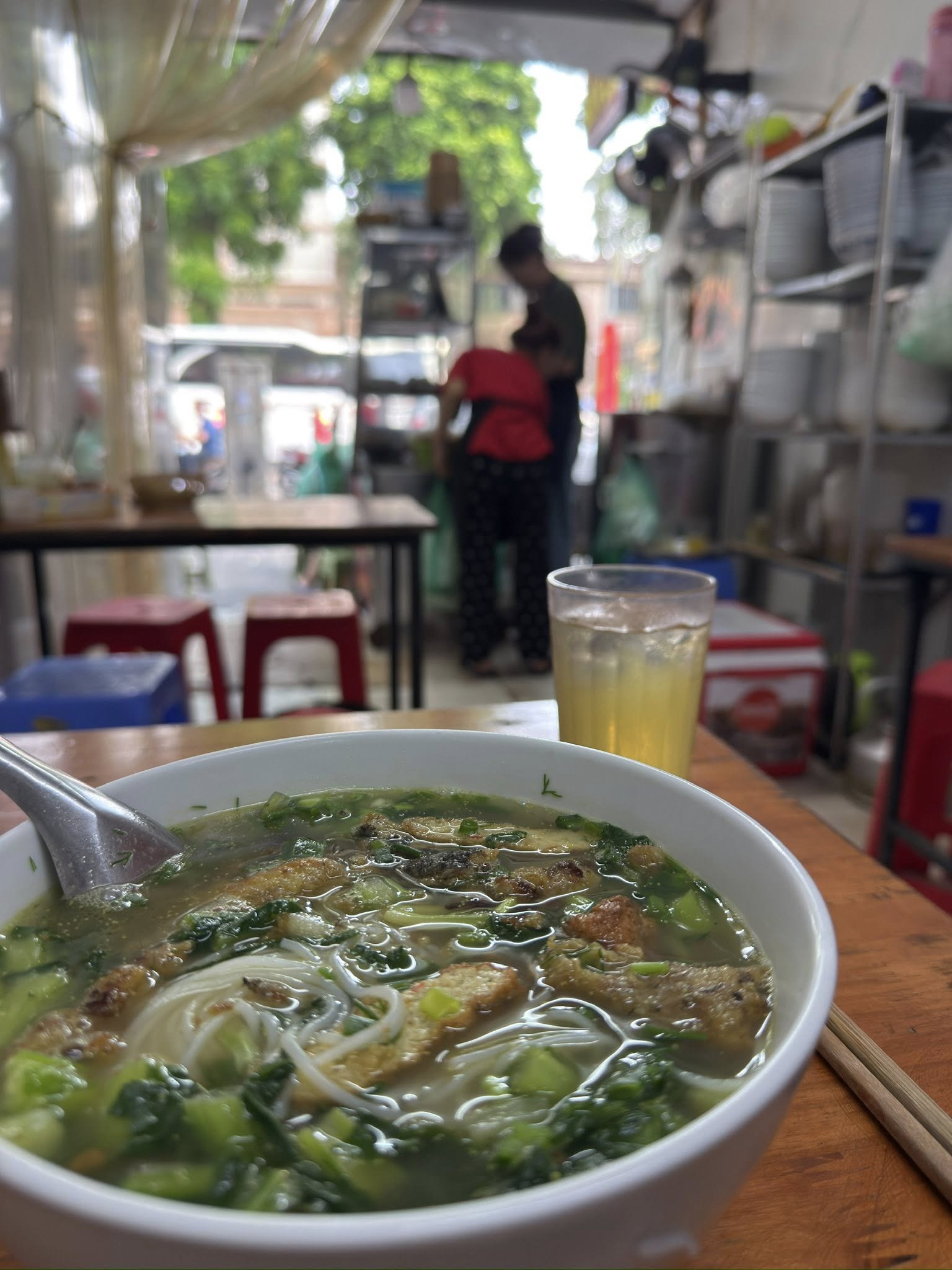 河內街邊小店裡的一碗興安魚肉河粉，前景是熱騰騰的魚粉與冰茶，背景是店家在檯前忙碌的身影。