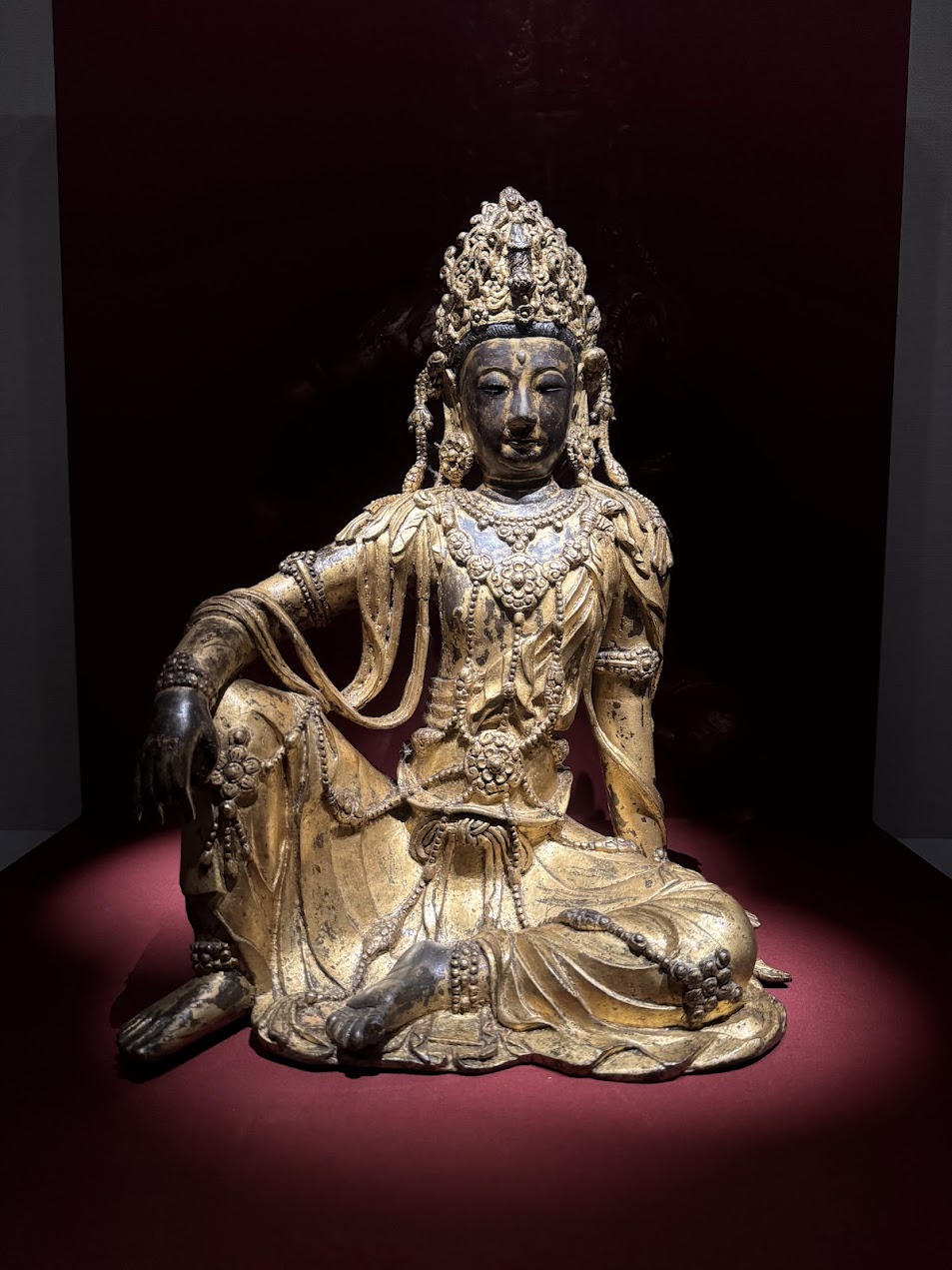 越南河內國家美術博物館展出的木雕漆金觀音菩薩坐像。佛像呈遊戲坐姿（Lalitasana），姿態自在優雅，面容慈祥靜謐。頭戴精緻繁複的寶冠，身披華麗瓔珞與飄逸天衣，展現出越南傳統佛教造像工藝中細膩的雕刻技法與莊嚴美感。