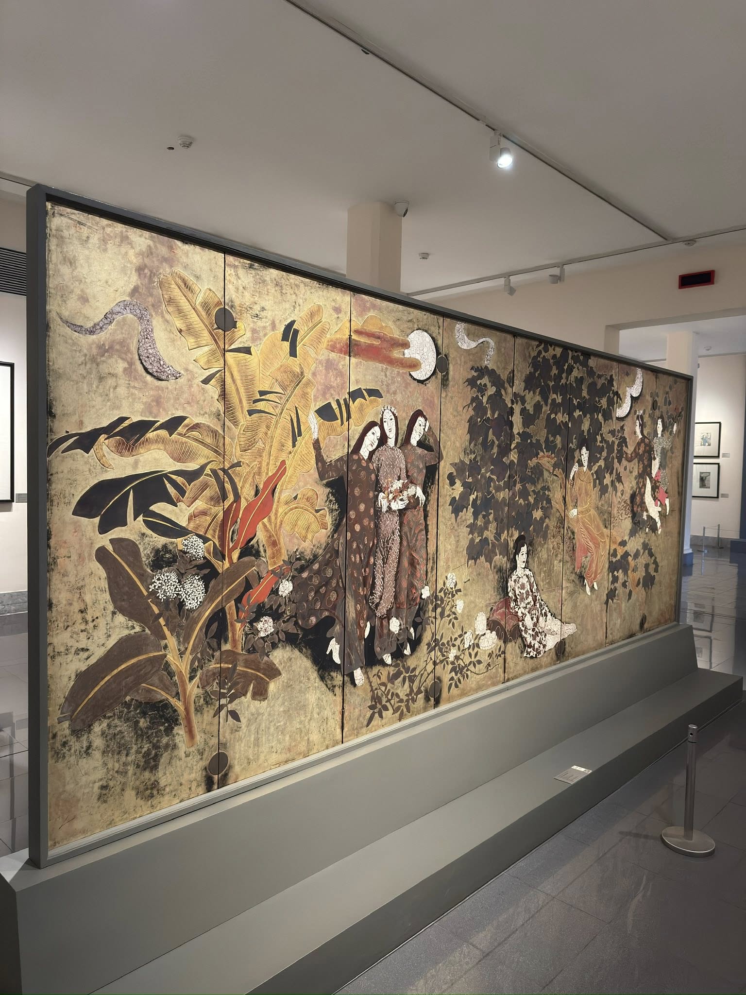 越南國家美術館展出的阮嘉智〈園中的少女〉全景漆畫，少女們站在蕉葉與庭園之間，以大片平面色塊與金色光澤呈現的長屏風作品