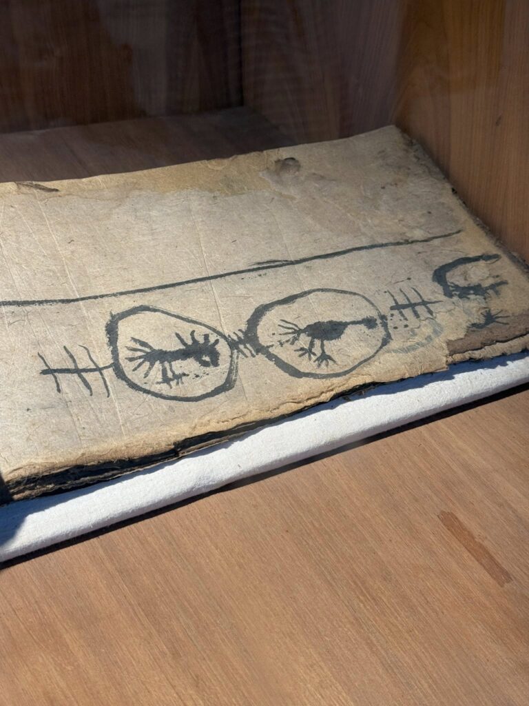 展示於博物館玻璃櫃中的古老紙本畫作，描繪雙重附體符號圖像，人物以符線連結形成「神靈通道」構圖，呈現東亞薩滿與道教巫儀中身體作為神靈媒介的象徵概念。