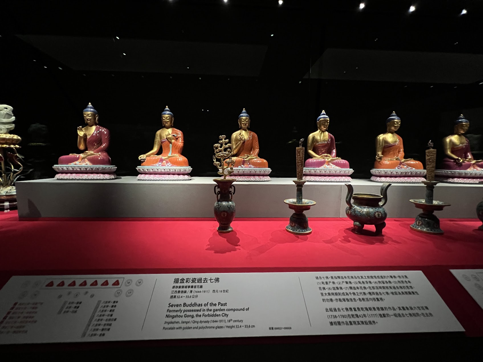 「北京故宮寧壽宮舊藏〈描金彩瓷過去七佛〉展件。七尊佛像以粉彩瓷呈現，面容柔和、衣紋簡潔，色彩介於玫瑰紅、赭橙與金色之間。佛像按傳統‘過去七佛’次序排列於展櫃中央，前方陳列清代宮廷祭器，如瓷質供瓶、燭台與爐鼎，反映清宮內廷將佛教造像、瓷器工藝與禮儀系統結合的美學。」
