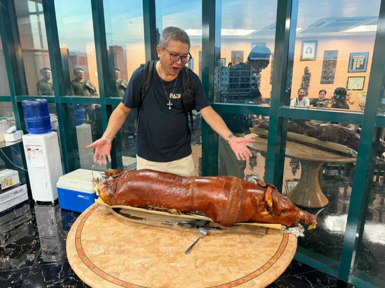 周端政在宿霧室內宴會空間與一整隻 Lechon 烤豬合照，站在圓桌旁露出驚喜表情，背後可見玻璃隔間與正在用餐的親友。