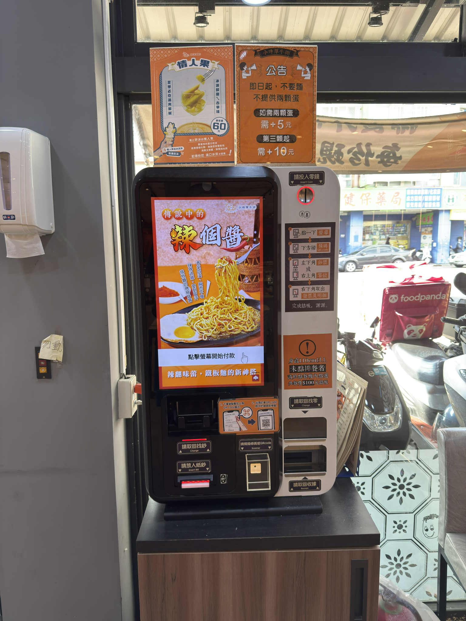 餐廳內的自助點餐與收款機，整合觸控螢幕、行動支付與現金投幣功能，用於取代人工櫃檯完成點餐與結帳流程，呈現服務流程自動化的現場場景。