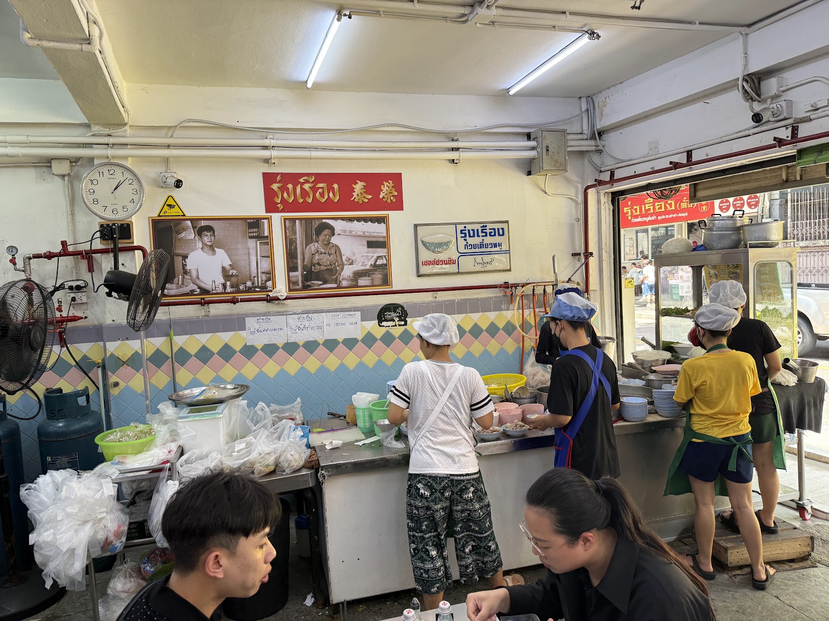 曼谷一間老字號豬肉湯麵店內景，師傅與客人在開放式廚台前忙碌，牆上掛著紅底招牌