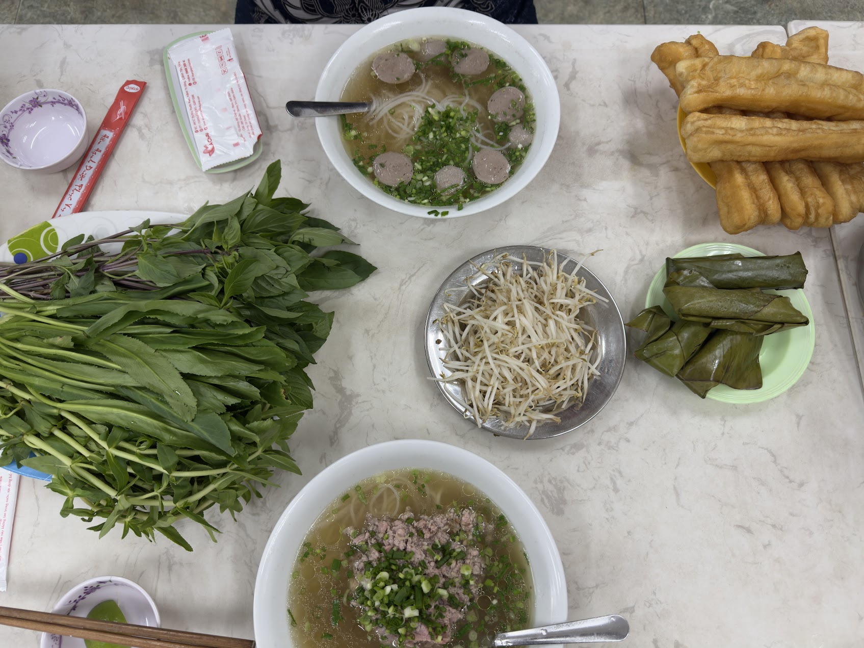 二つの米麺スープ、もやし、大量の香草、揚げパン、包み菓子が並ぶテーブル。東南アジアのスープ食が周辺の要素と一緒に組み立てられている様子。