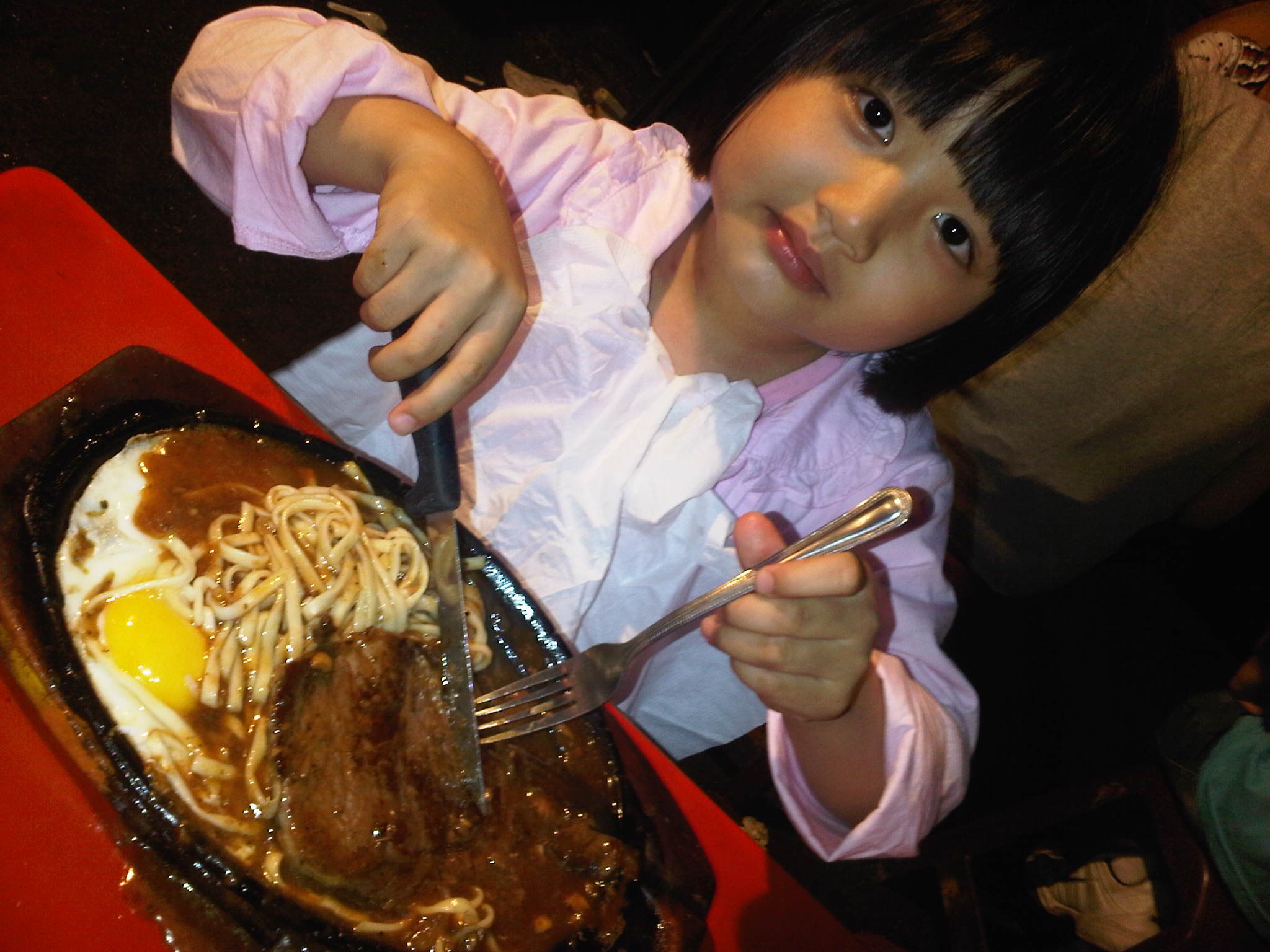 台湾式の鉄板ステーキセットの前に座る小さな女の子。麺、卵、ソースを含む一食が、家族の外食記憶になっていく様子を示している。