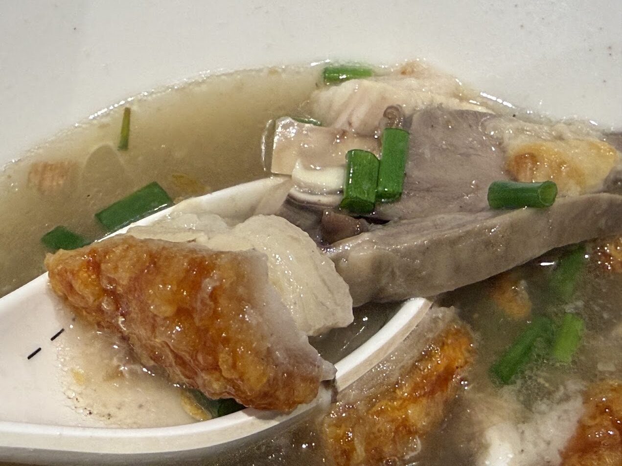 湯匙舀起一塊脆皮豬肉，碗中可見清湯、肉片與蔥花，呈現泰式湯麵對口感層次的重視
