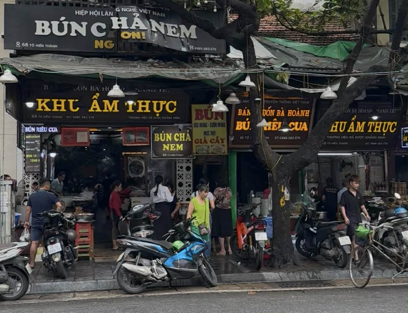 ハノイのバインクオン老舗の昼時の店先。バイクが並び、店内外に客の出入りが続き、地元の人の日常の昼食風景がそのまま立ち上がっている。