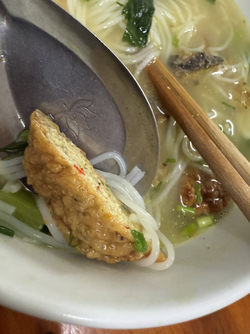 A close-up spoonful of fried fish in Canh cá rô Hưng Yên, with rice noodles, greens, and broth visible around it.