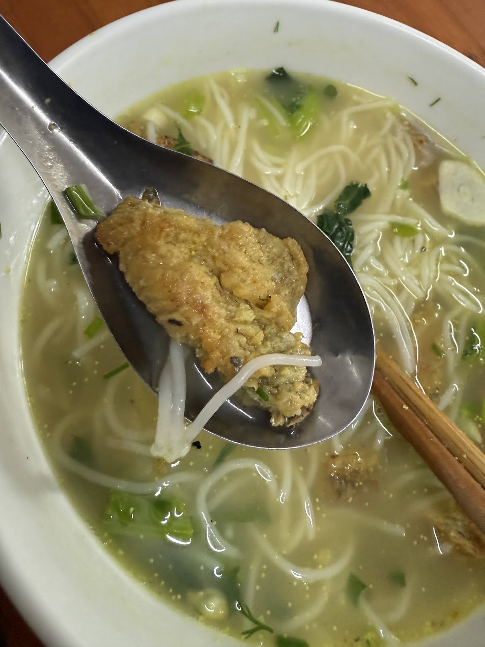 湯匙舀起河內 Canh cá rô Hưng Yên 中的炸魚塊,背景可見清亮魚湯、白色米麵與青綠蔬菜。