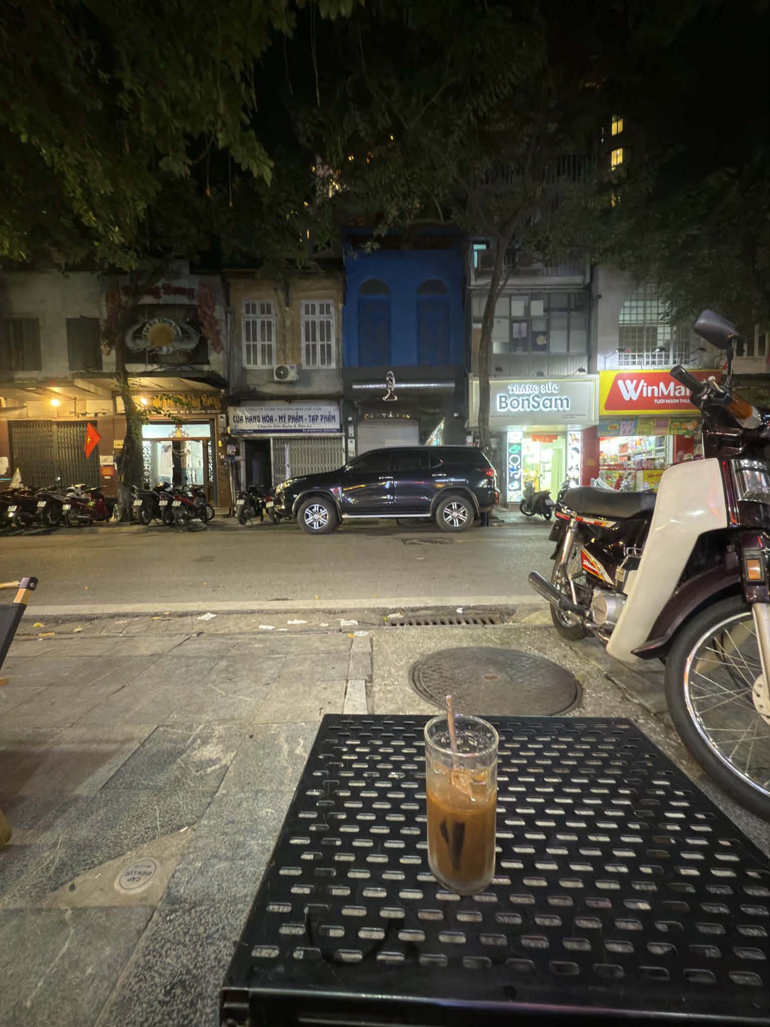 ハノイの夜、街角のテーブルに置かれたアイスコーヒー。近くにはバイクが停まり、向かいの店にはまだ灯りが残っていて、都市の静かな余韻が感じられる。