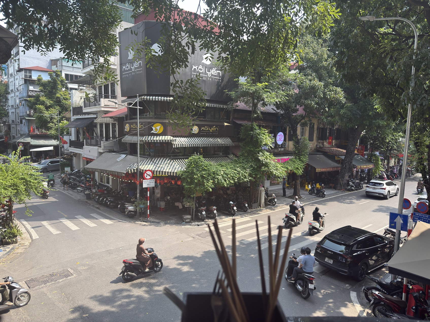 ハノイの朝の街角。木陰、交差点、バイク、店先が重なり、路上の朝食が置かれている都市の本当の日常リズムが見える。