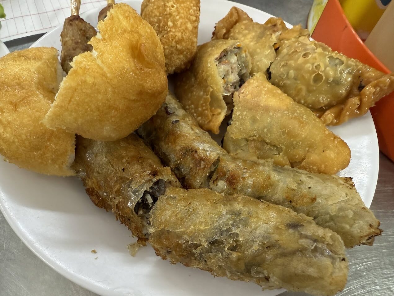 ハノイの街角の朝食で出された揚げ物の盛り合わせ。揚げ春巻き、揚げ饅頭、肉棒、揚げ小点が並び、庶民的で温かな朝の一皿になっている。