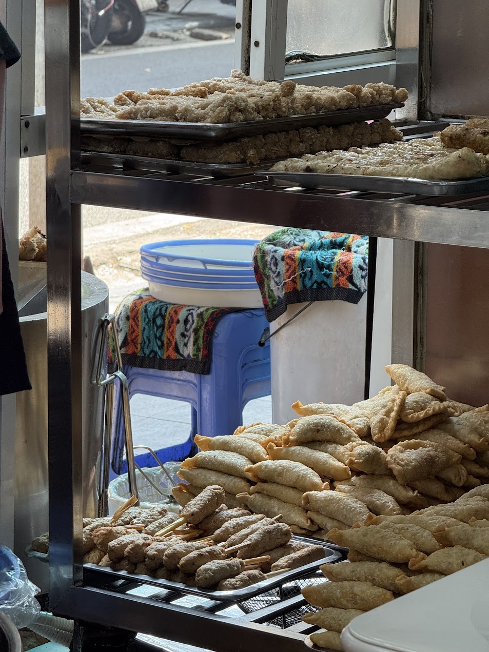 ハノイの街角の朝食屋台で、揚げ春巻き、揚げ饅頭、肉串などの朝の揚げ物が整然と積み上げられた鉄棚の前場風景。