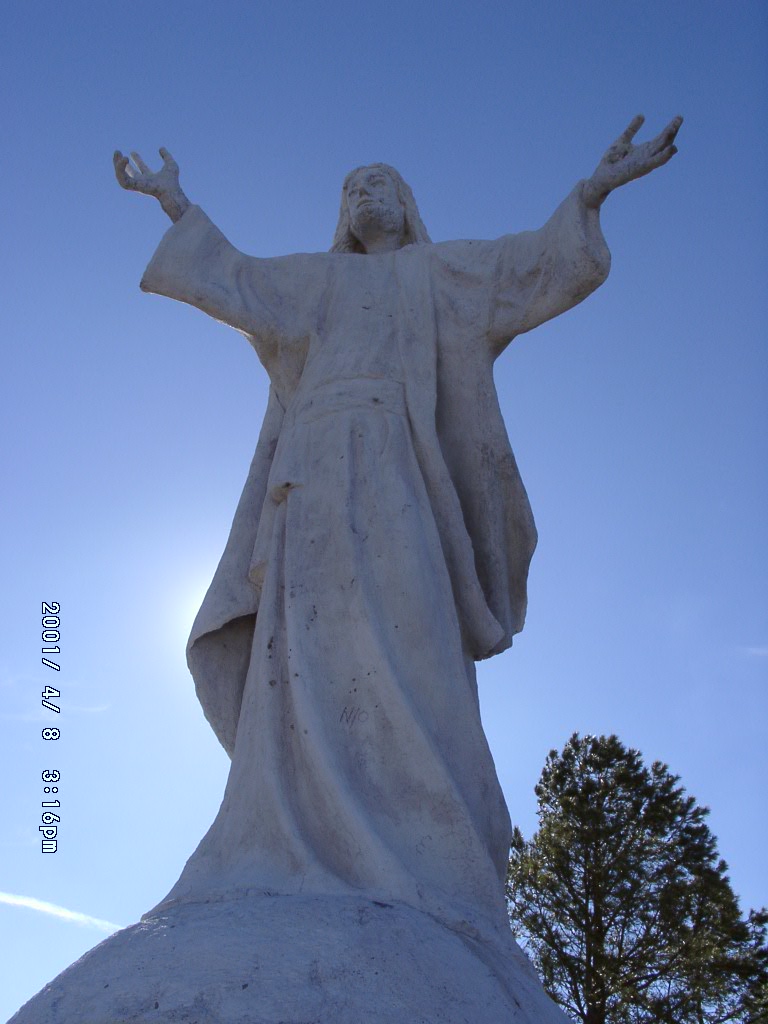 White Christ statue with open arms in Mexico, standing against sky and upland landscape, showing how sacred imagery in Latin America grows into local topography and public space.
