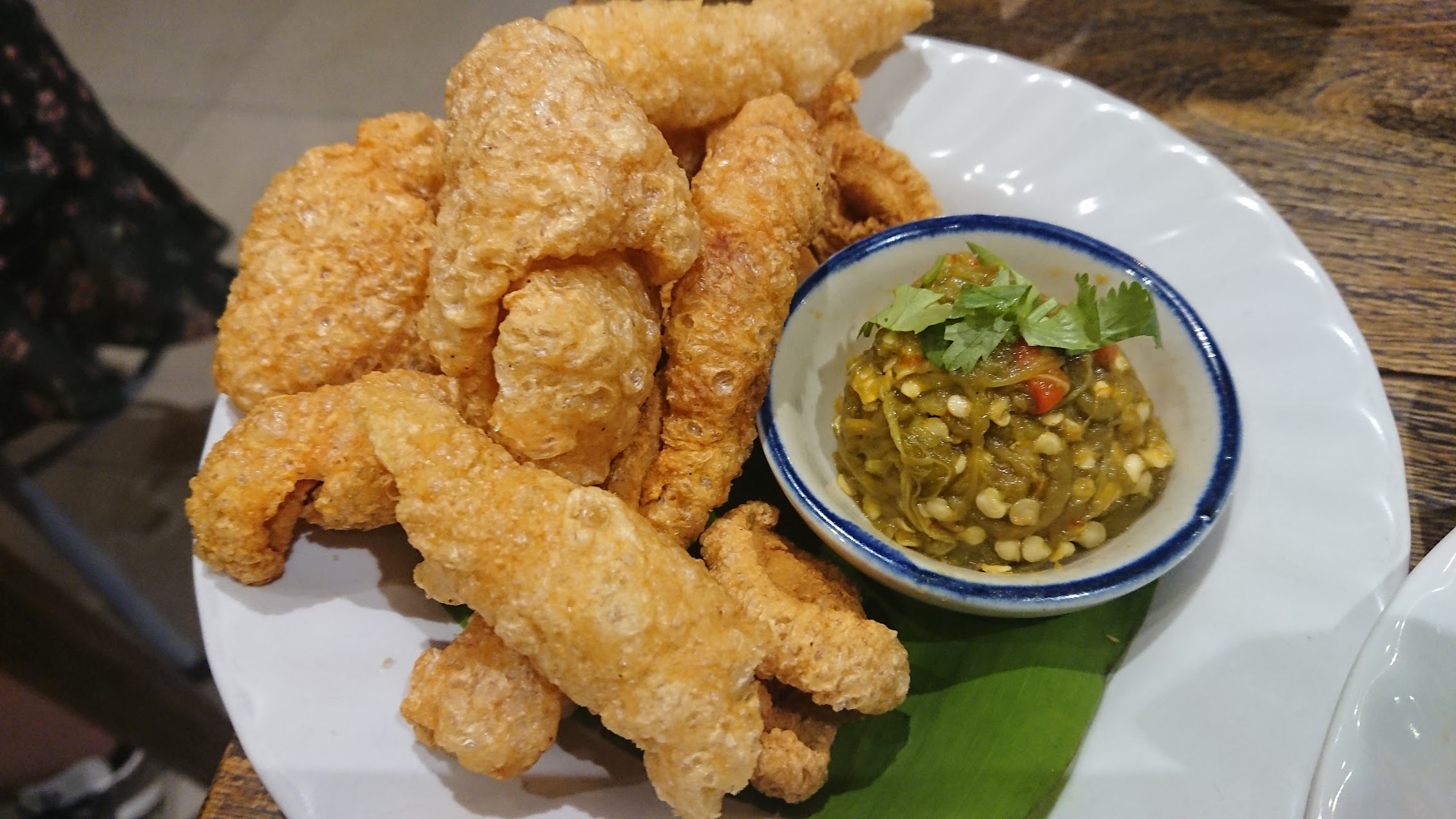 青唐辛子のディップを添えて供されるタイの豚皮揚げ。袋菓子ではなく、日常の食卓の一部として食べられていることがわかる。